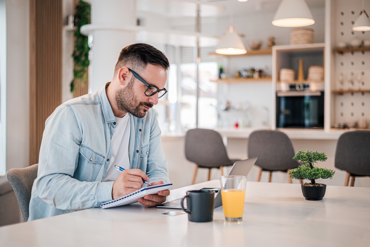 Man working on a business plan at the kitchen table with a notepad and laptop.